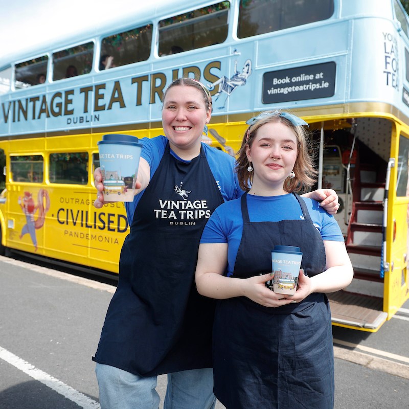 Two Vintage Tea Trips staff members in uniform holding the branded travel mugs in front of the bus