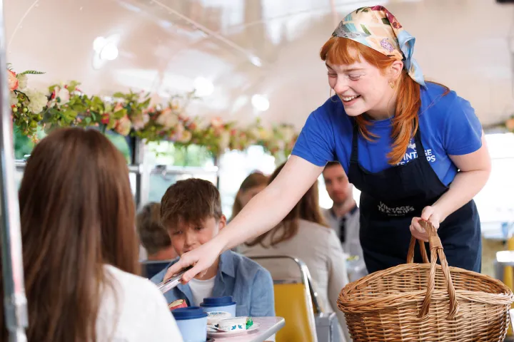 Smiling server with basket on Vintage Tea Trips, offering a scone to seated customers.