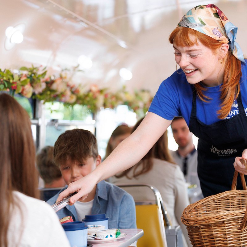 Smiling server with basket on Vintage Tea Trips, offering a scone to seated customers.