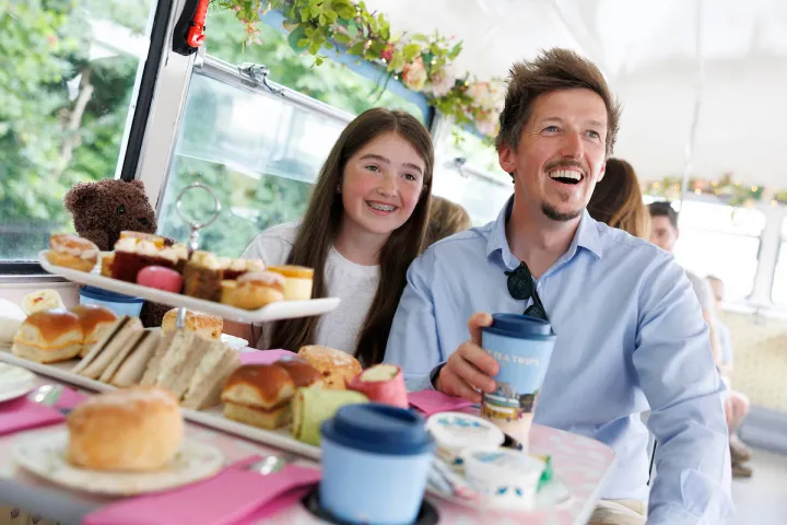 Man and girl smiling on bus with tea and pastries on table.