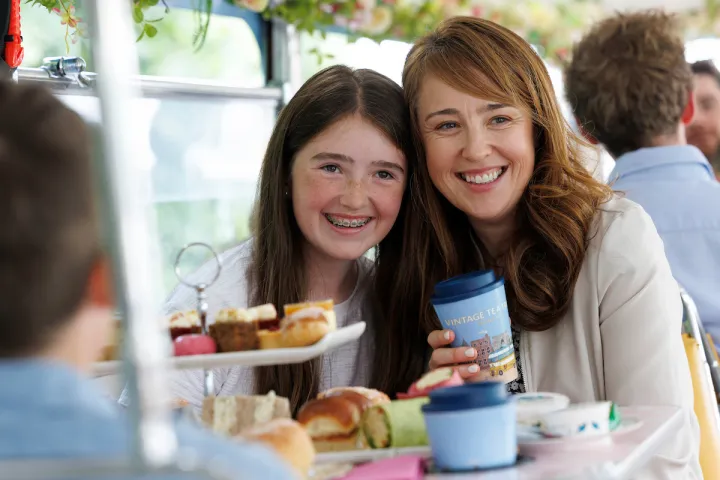 Smiling woman and girl with food on a table, holding a cup.