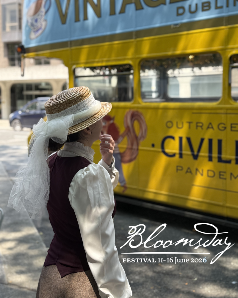 Person in vintage attire stands by a yellow bus with 'Bloomsday Festival 11-16 June 2026' text.