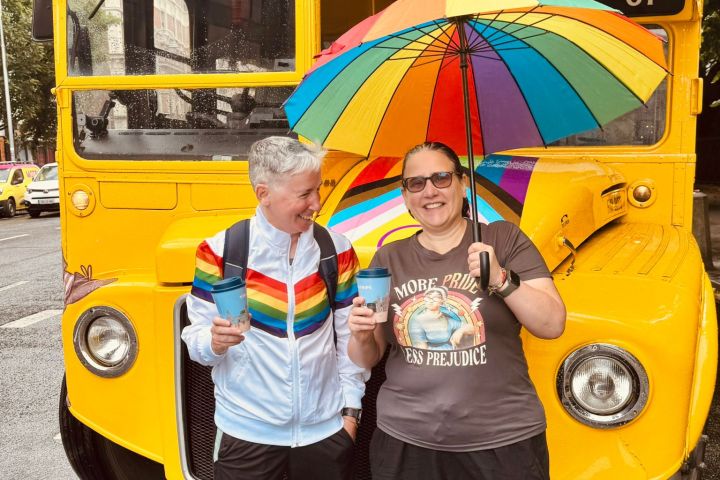 Two people with a rainbow umbrella stand in front of a vintage yellow bus.