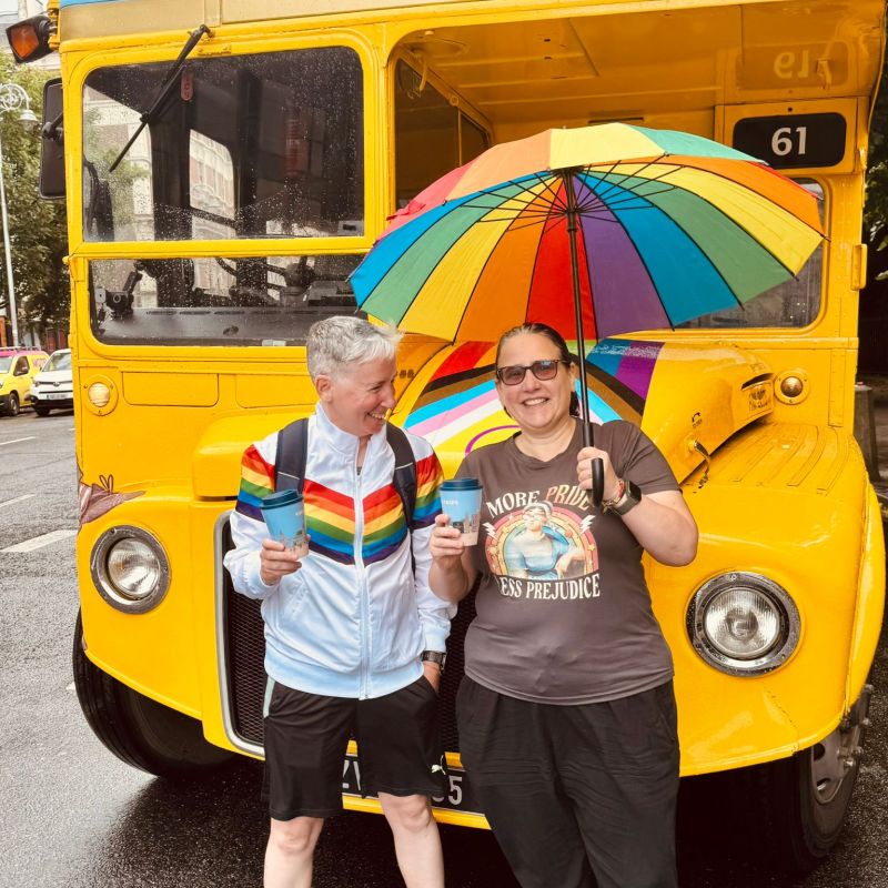 Two people with a rainbow umbrella stand in front of a vintage yellow bus.