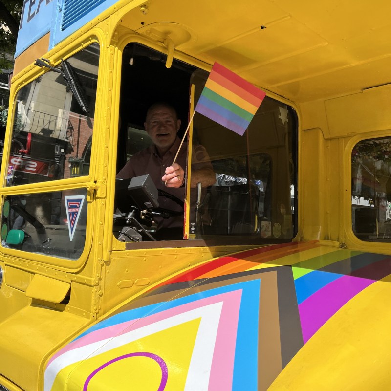 Bus driver waving Pride flag out of the Vintage Tea Trips bus window