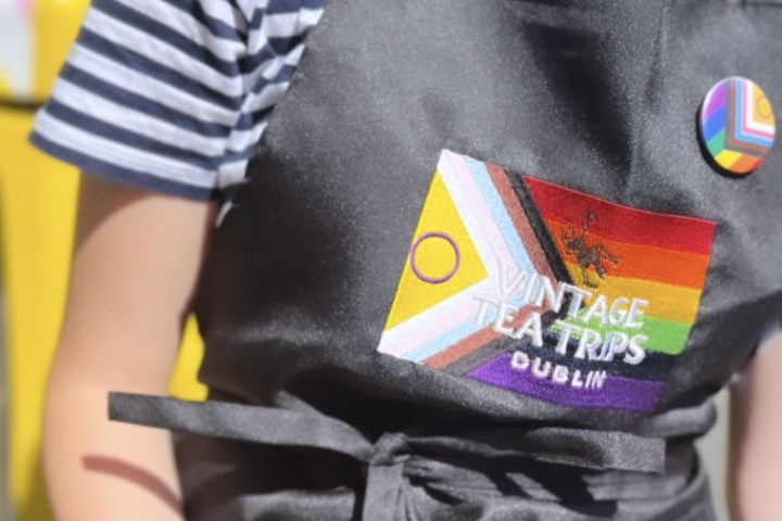 Close up of person wearing an apron with a Pride badge and a Pride flag behind the Vintage Tea Trips logo