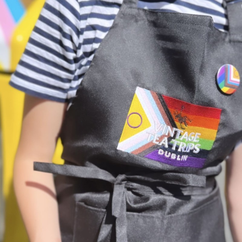 Close up of person wearing an apron with a Pride badge and a Pride flag behind the Vintage Tea Trips logo