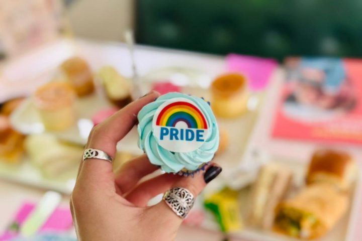 Hand holding cupcake with rainbow 'PRIDE' frosting above table with food.