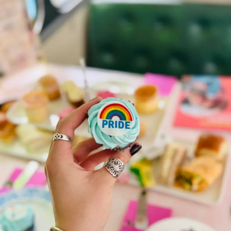 Hand holding cupcake with rainbow 'PRIDE' frosting above table with food.