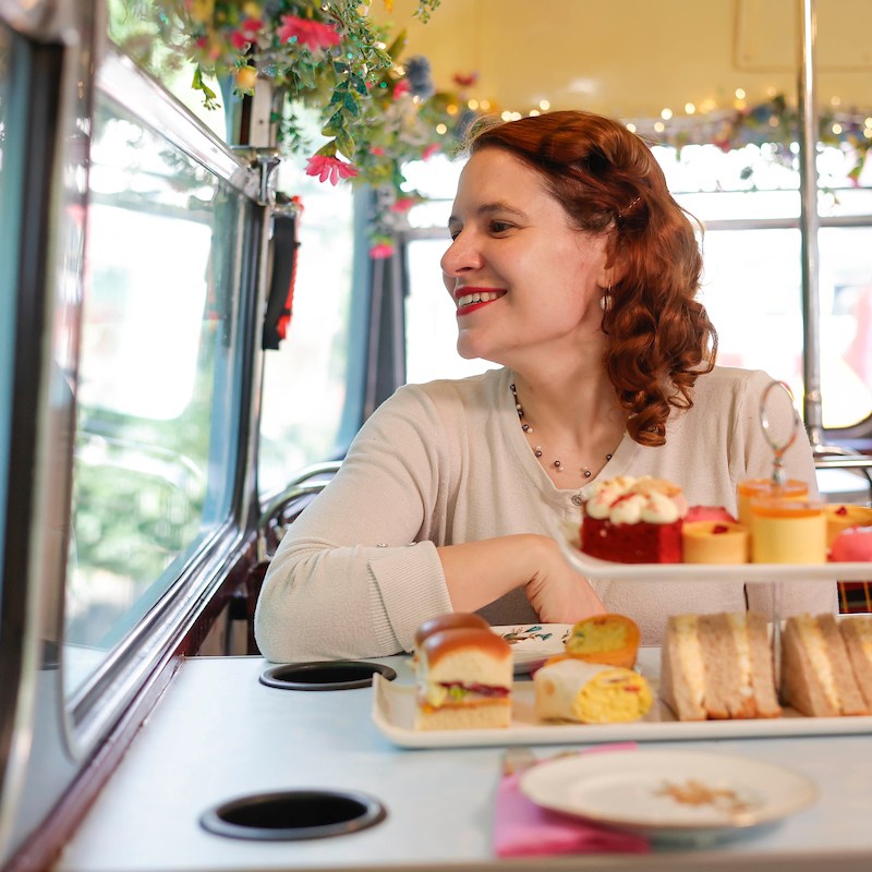 Woman smiling out of bus window with arms crossed and afternoon tea on the table in front of her