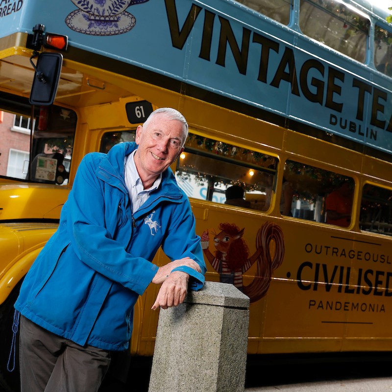 Bus driver in blue jacket resting arm on a concrete bollard with the Vintage Tea Trips bus behind him