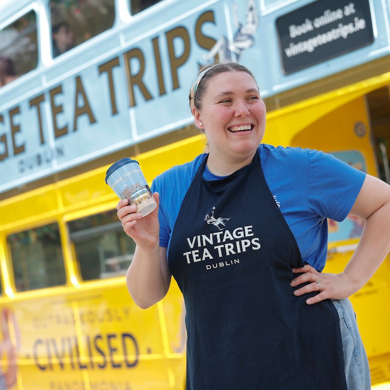 Smiling woman wearing Vintage Tea Trips uniform holding a travel mug in one hand and the other on her hip