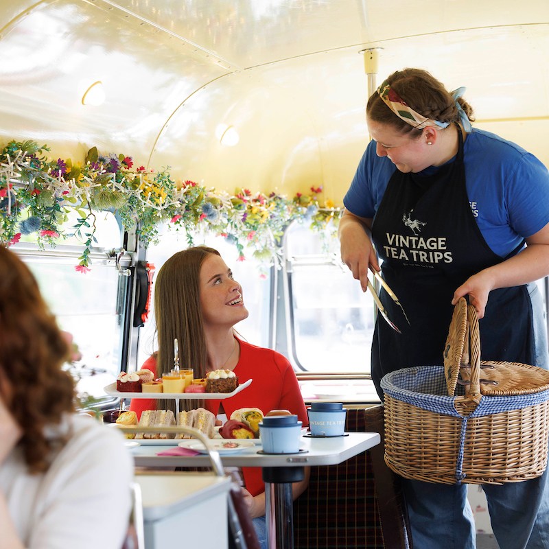 Vintage Tea Trips staff member holding a wicket basket and tongs while talking to bus passenger