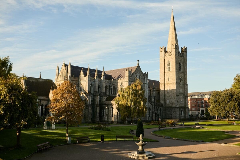 Stone cathedral with tall spire, surrounded by trees and a pathway in a sunny park.