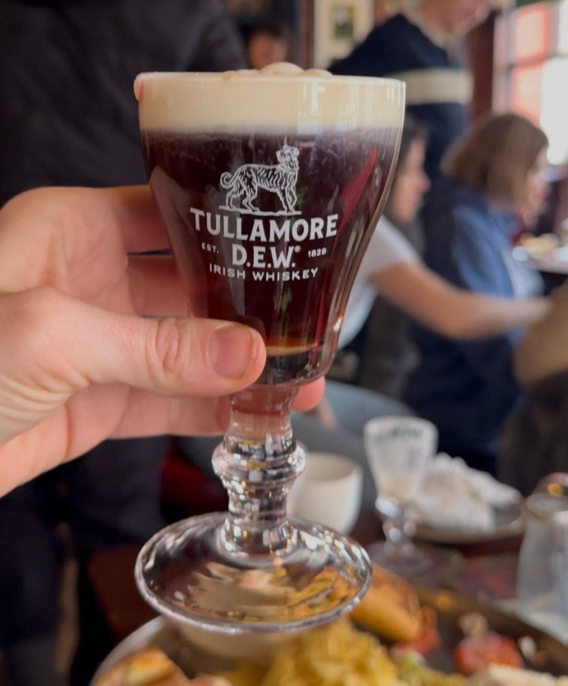 Hand holding an Irish whiskey glass with a drink, people in background.