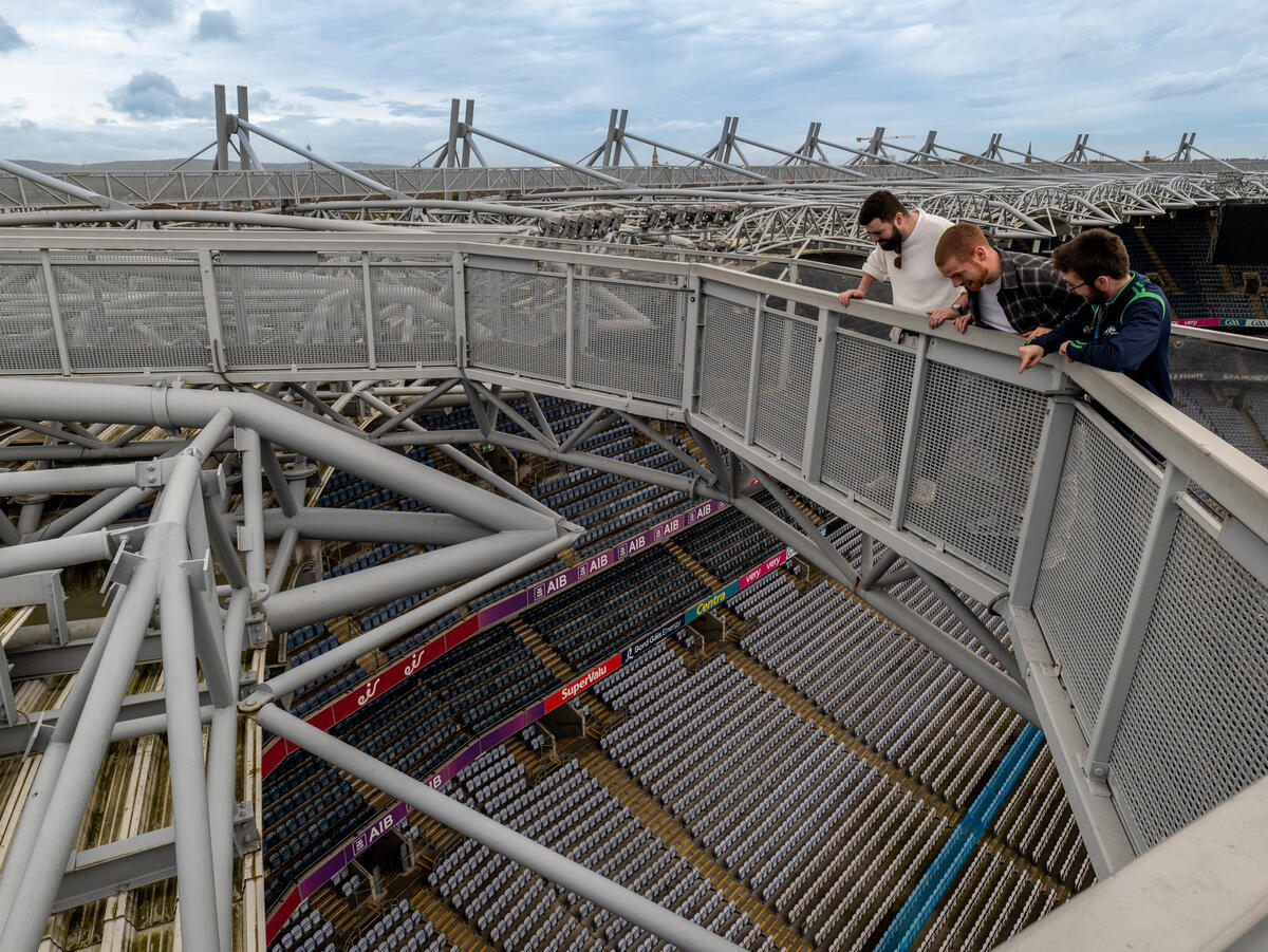 Three men look down from a stadium rooftop walkway with metal beams and empty seats below.