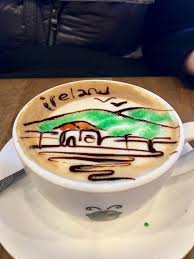 Cappuccino with Ireland-themed art in froth, depicting a landscape and text, in a white cup on a saucer.