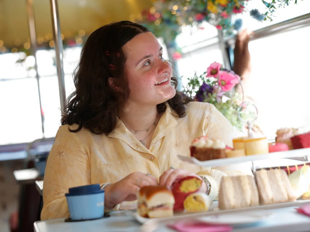 Person smiling at table with sandwiches, pastries, and flowers, inside a bright setting.