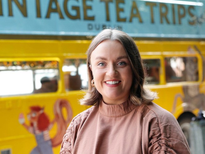 Smiling woman in front of a vintage yellow bus with 'Tea Trips' sign.