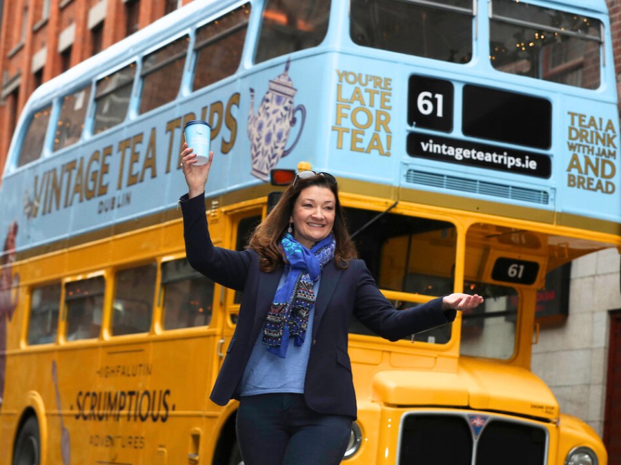Smiling person holds cup in front of a vintage double-decker bus with tea-themed decorations.