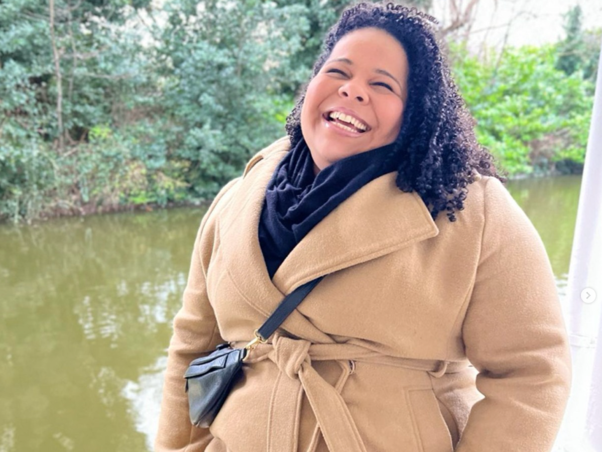 Smiling person in a tan coat stands near a river with trees in the background.