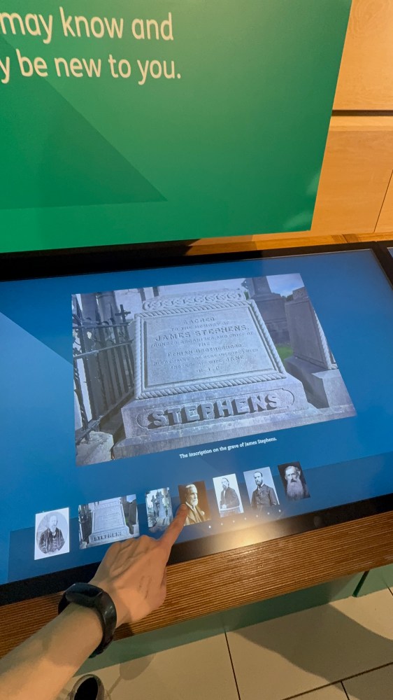 Person pointing at a touchscreen display showing a gravestone image.