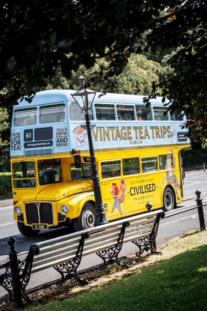 Yellow vintage double-decker bus on a street with 'Vintage Tea Trips' ad, surrounded by trees.