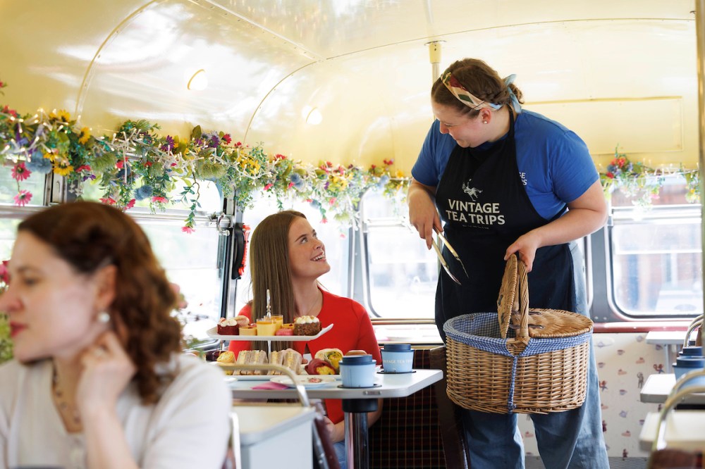 Server with apron offers a basket to seated woman in a decorated bus cafe.