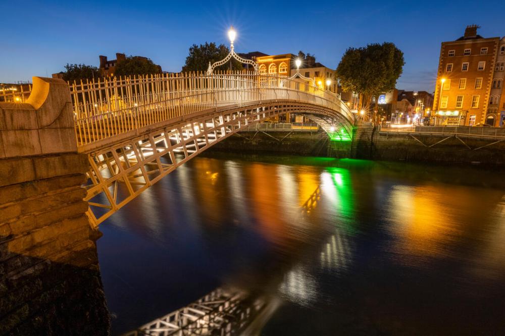 Arched footbridge over river with city lights reflecting on water at dusk.