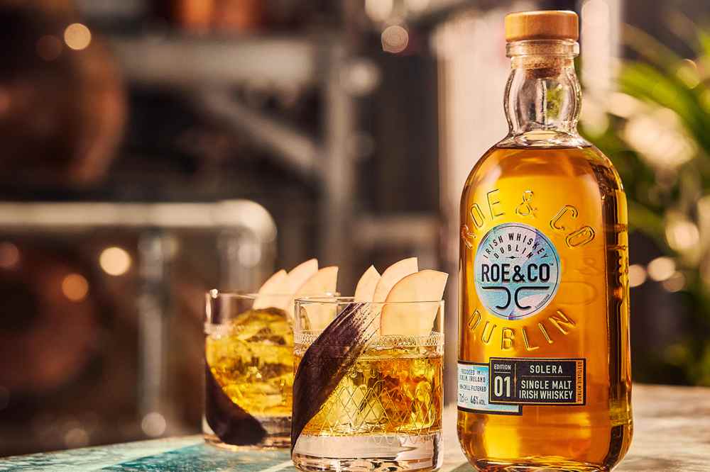 Bottle of Roe & Co whiskey with two glasses of whiskey and apple slices.