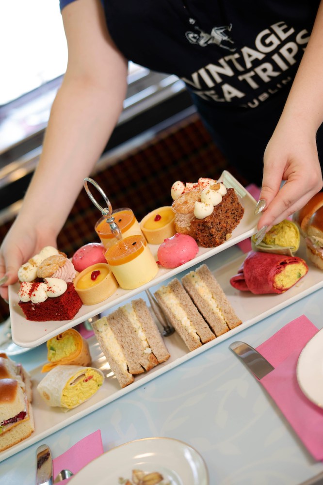 Person serving a variety of pastries and sandwiches on a tea tray.