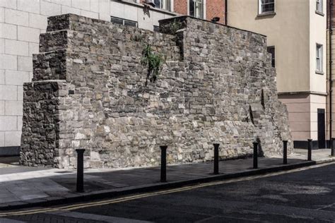 Old stone wall beside a city sidewalk with bollards.