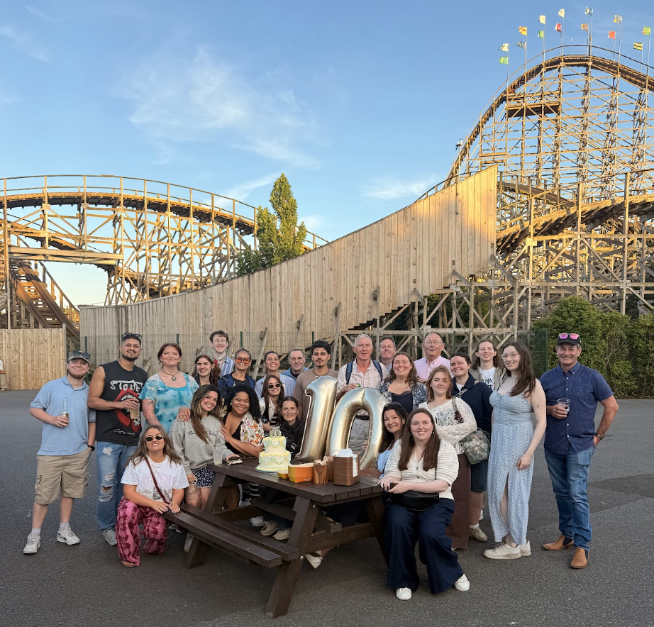 Group of people at picnic table in front of wooden roller coaster.
