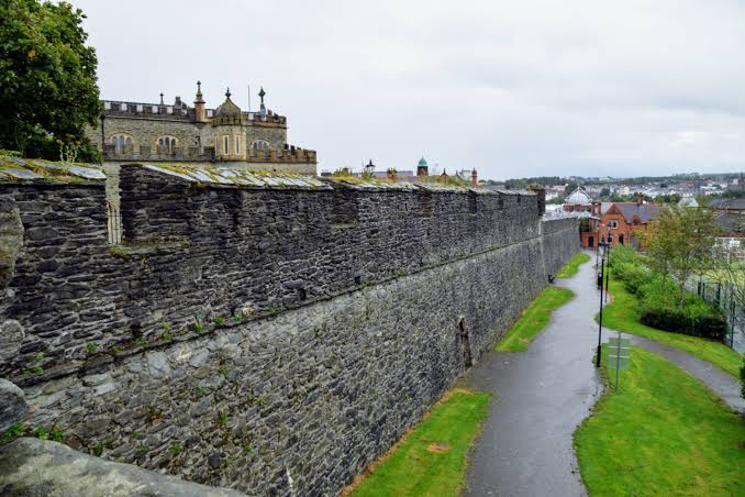 Stone wall with adjacent path and distant buildings on a cloudy day.