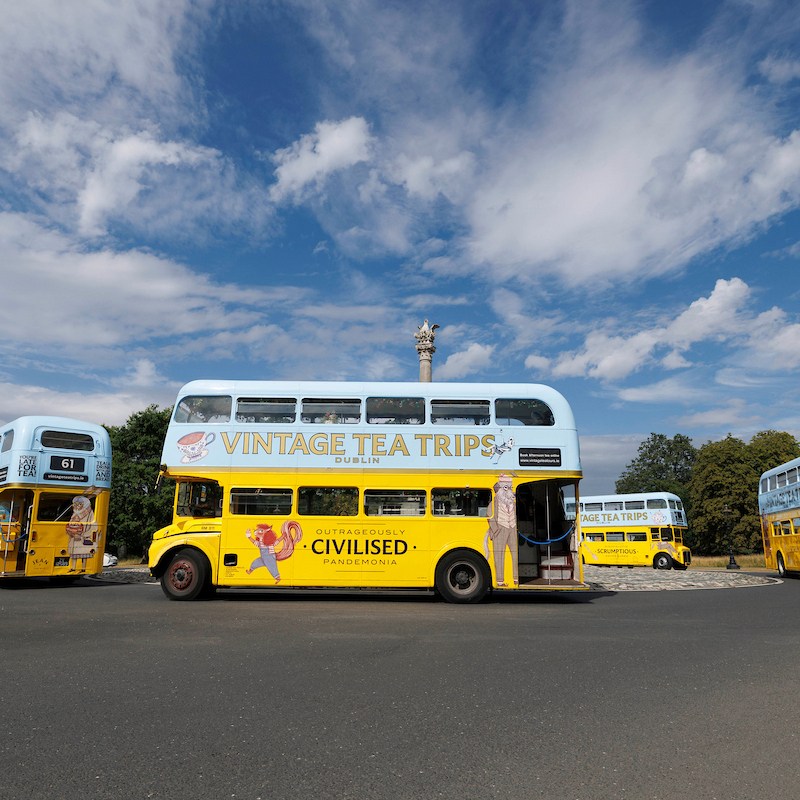 Four vintage yellow double-decker buses driving around a roundabout in Phoenix Park under a blue cloudy sky