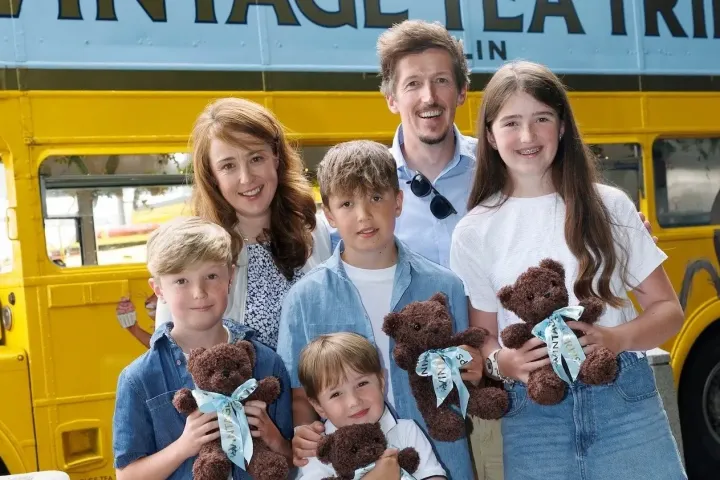 Family with children holding teddy bears in front of a yellow vintage bus.