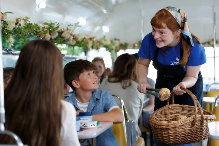 Waitress serves bread to kids on a bus with floral decor.
