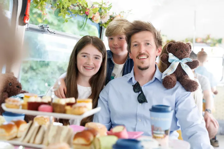 Family enjoying tea on a bus with pastries, flowers, and teddy bears.