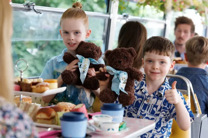 Two children hold teddy bears on a bus with food in the foreground.