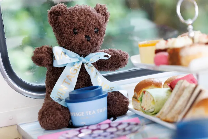 A teddy bear with a blue ribbon beside a tray of afternoon tea snacks on a table.
