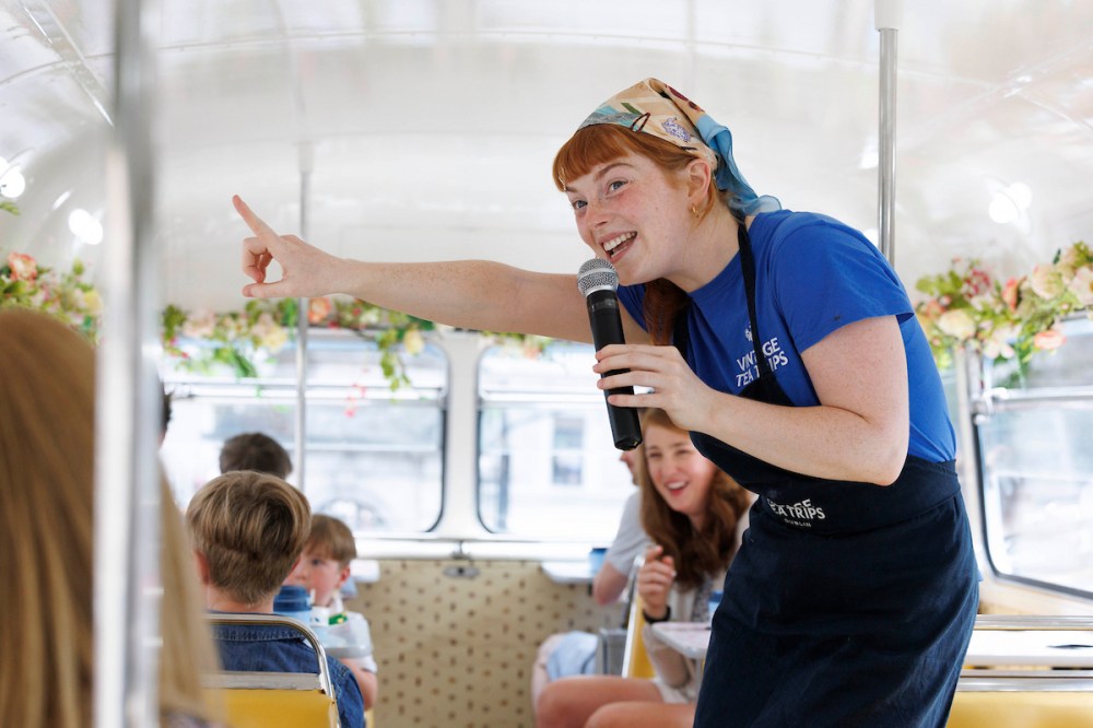 Smiling woman with microphone entertains children on floral-decorated bus.