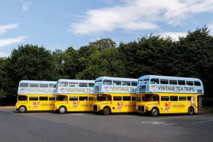 Four yellow vintage double-decker buses parked in a row near trees under a blue sky.