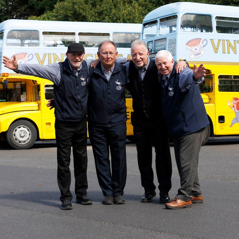 Four smiling bus drivers in front of vintage double-decker buses.